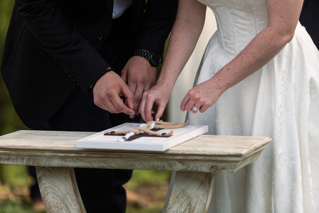 Close-up of the couple’s hands during a unity ceremony, carefully tying a knot with a thick rope on a wooden table, symbolizing their marriage. The bride’s white gown and the groom’s dark suit frame the intimate moment.