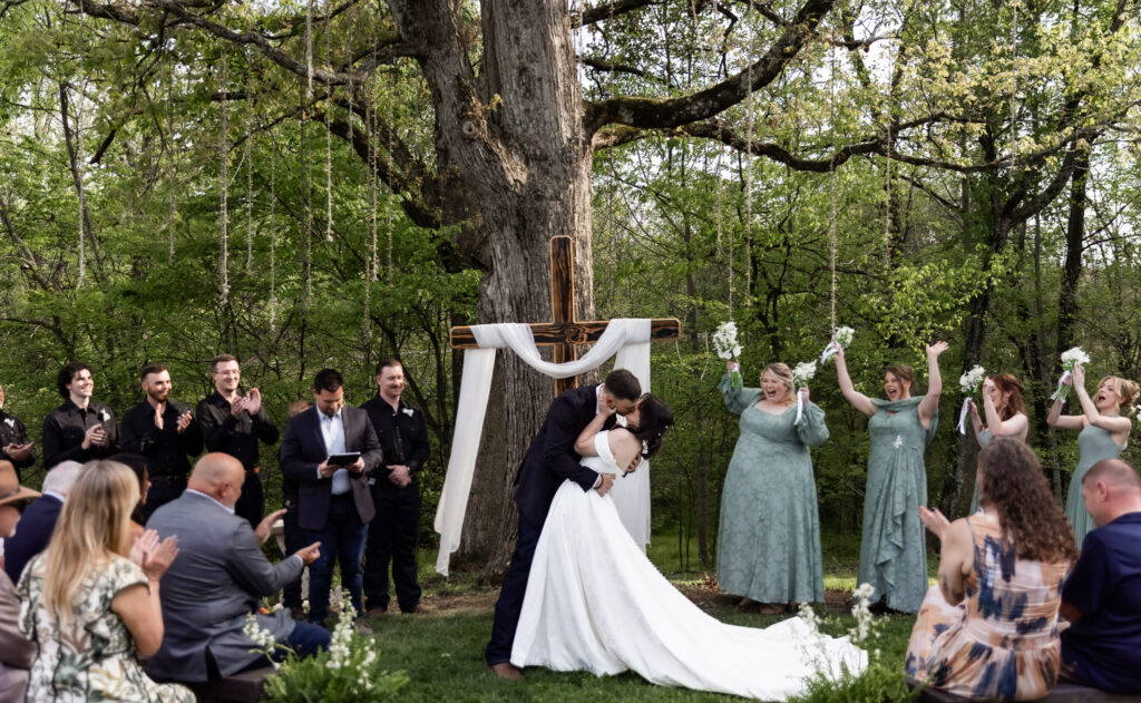 The couple shares their first kiss beneath a large wooden cross draped in soft white fabric during an outdoor Greenwood Oaks wedding ceremony, surrounded by towering trees and lush greenery, while guests seated on wooden benches clap and smile and bridesmaids in sage green dresses throw their bouquets up in celebration.