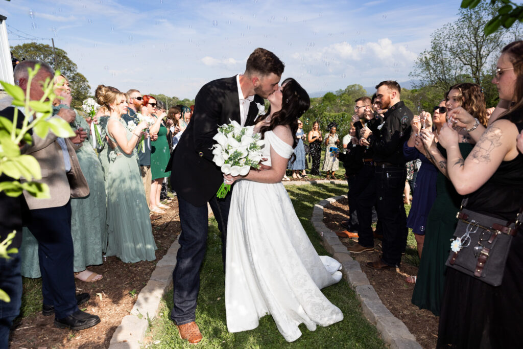 The bride and groom kiss at the end of a curved stone-lined aisle during their recessional at a Greenwood Oaks wedding, the bride and groom kiss again as guests on both sides blow bubbles into the air, sunlight catching each floating sphere while the bride holds a bouquet of white flowers and her dress trails behind her.