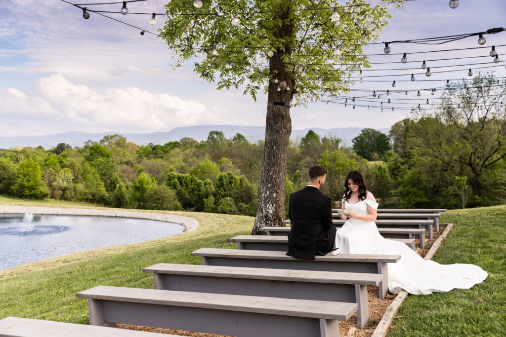Seated side by side on simple wooden ceremony benches at a Greenwood Oaks wedding, the couple faces each other quietly as the bride reads her vows from a small booklet, with a pond, distant mountains, and string lights hanging overhead creating a peaceful, open-air setting.