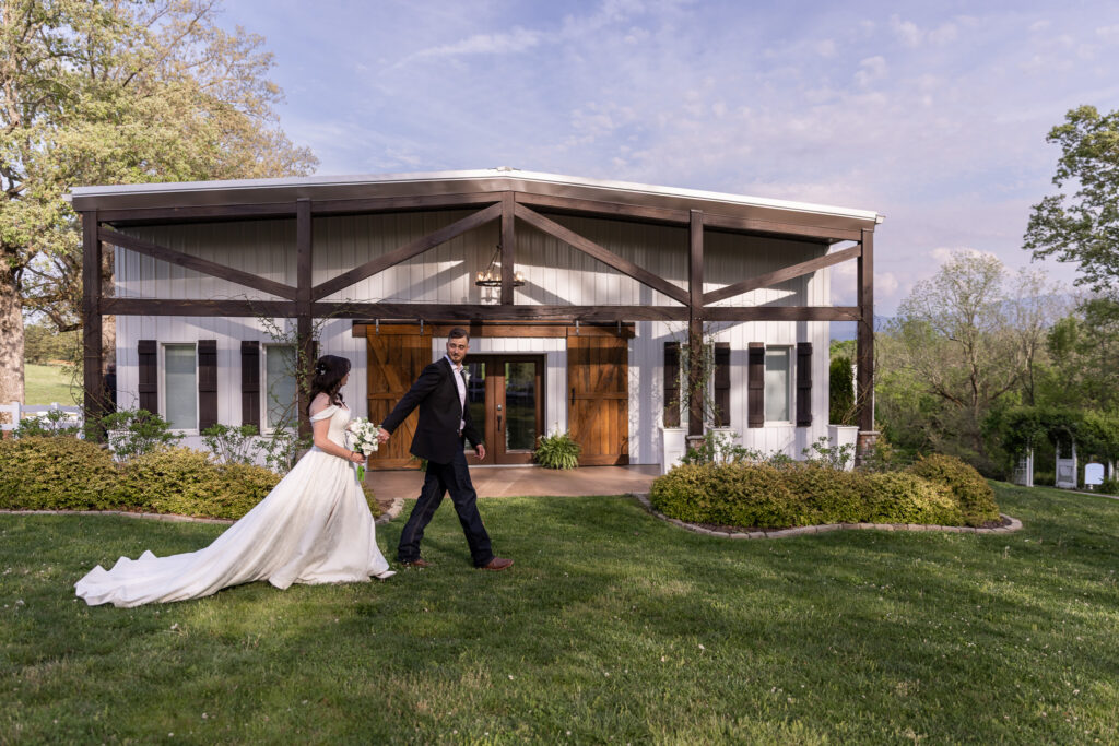 In front of a modern barn-style building at a Greenwood Oaks wedding, the groom leads the bride by the hand across a green lawn, glancing back at her as her long train stretches behind and wooden beams and large barn doors frame the entrance in the background.