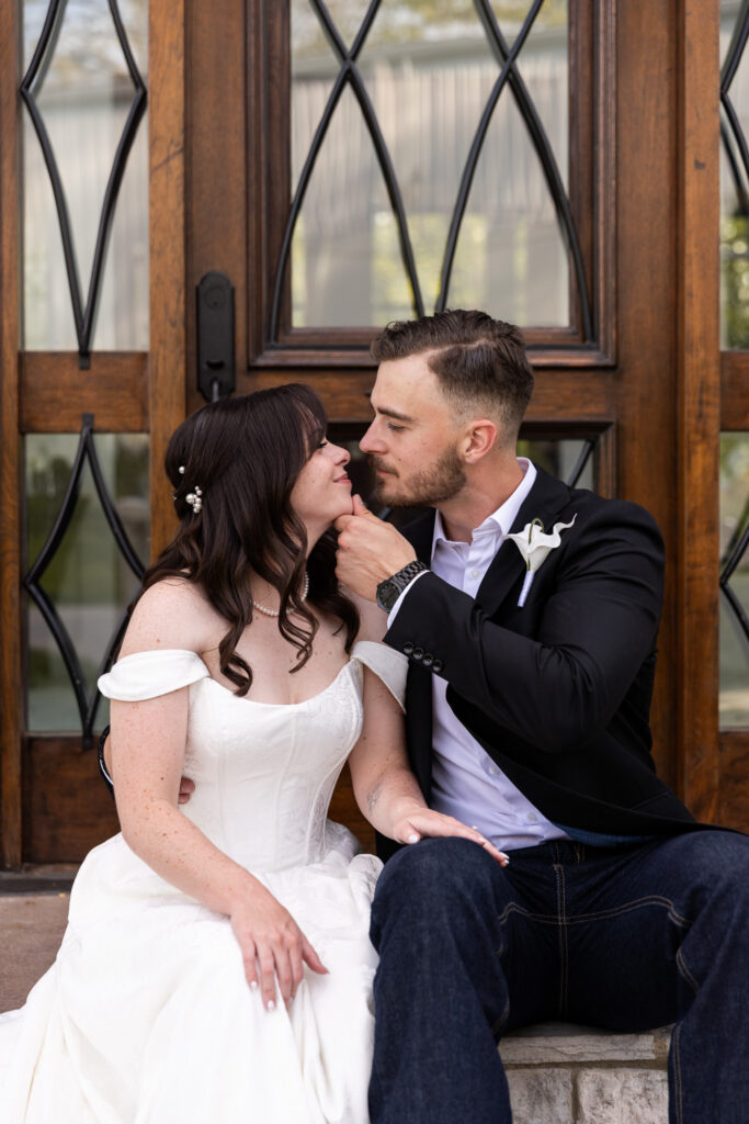 Intimate portrait of the bride and groom seated together in front of a wooden door, leaning in close with soft smiles. The groom lifts the bride’s chin slightly as they share a quiet, affectionate moment before their ceremony.