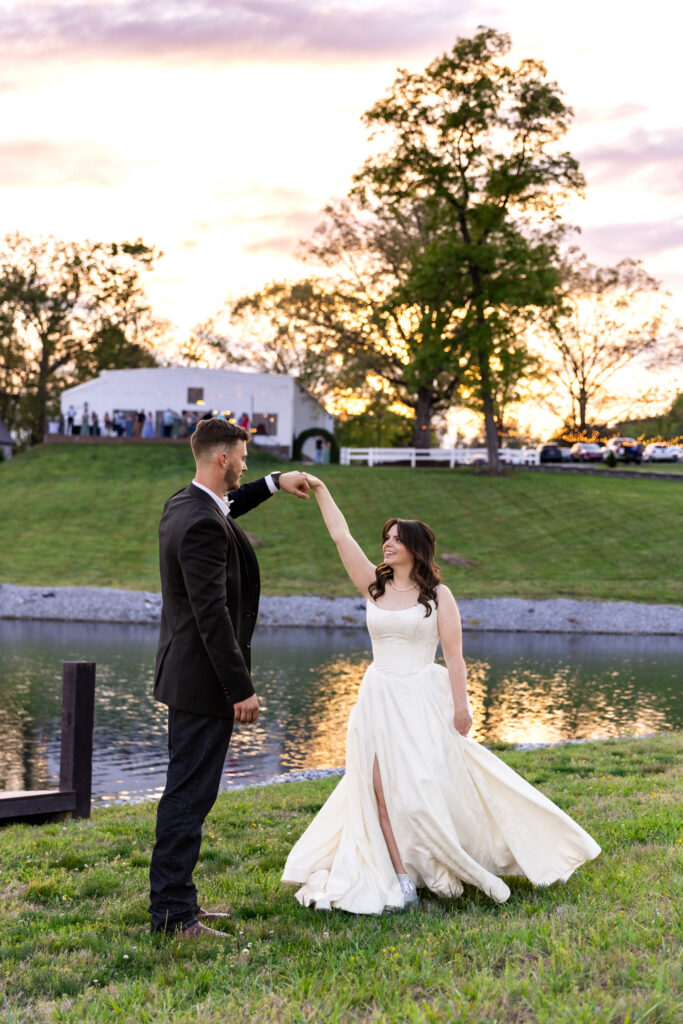 Near the edge of a pond at sunset during a Greenwood Oaks wedding, the groom lifts the bride’s hand as she twirls beneath it, her dress flowing in motion while golden light reflects off the water and a softly lit building sits in the distance.