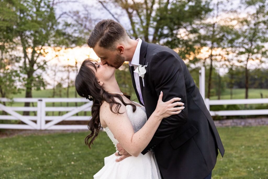 Romantic portrait of the bride and groom sharing a kiss outdoors at sunset, the bride dipping slightly in the groom’s arms. A white fence and soft greenery in the background frame the golden hour light.