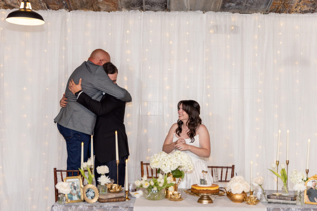 Reception moment where the groom embraces another guest in a heartfelt hug as the guest stands upon a chair to meet the grooms heights. The bride, seated nearby, smiles warmly as candles, white florals, framed photos, and a small cake decorate the table behind them.