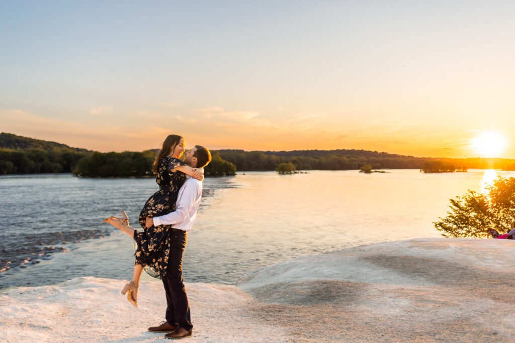 An engaged couple embraces on a rocky overlook during sunset at a White Cliffs of Conoy engagement session, with the glowing orange sun reflecting across the river behind them. The man lifts his fiancée into the air while she smiles down at him with her arms around his shoulders, her floral dress flowing softly in the breeze. The wide landscape emphasizes the open water, distant islands of trees, and expansive evening sky.