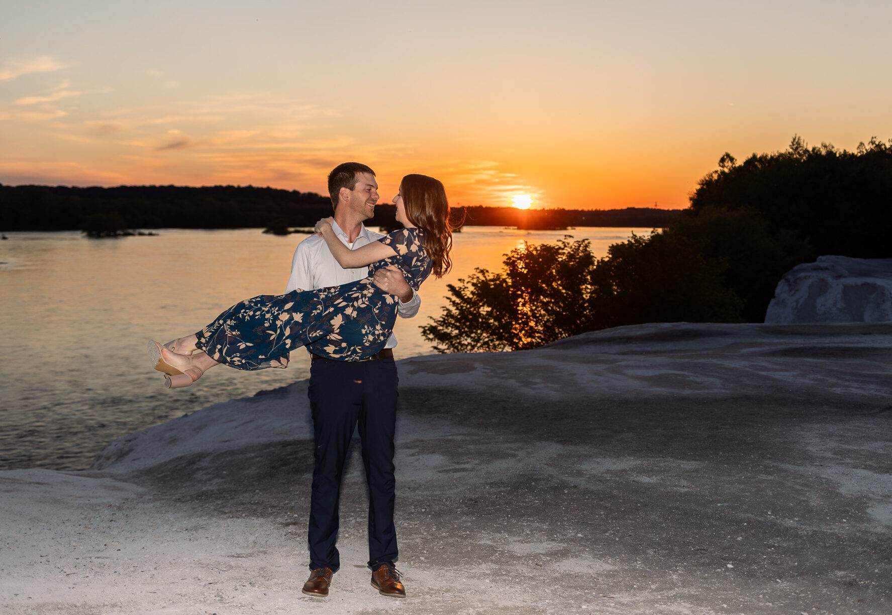 A man cradles his fiancée in his arms during sunset at a White Cliffs of Conoy engagement session, standing on the edge of a dramatic rocky overlook above the river. The couple smiles at one another while the vivid orange sun sinks low behind them, silhouetting the surrounding trees and reflecting warm light across the water. Her navy floral dress drapes across his arms while her nude heels extend outward toward the glowing horizon.