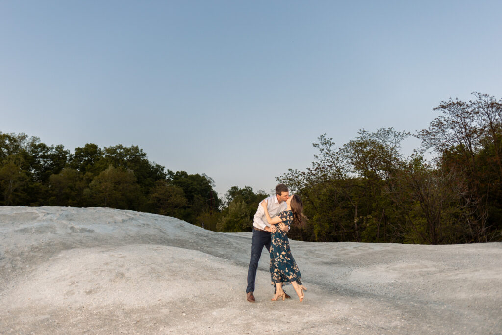 A couple shares a kiss while standing hand in hand atop the rolling white rock landscape during a White Cliffs of Conoy engagement session. The woman reaches up toward her fiancé’s face while he leans down to meet her kiss. The pale stone hills curve gently around them, framed by dense green trees and a fading blue evening sky in the background.