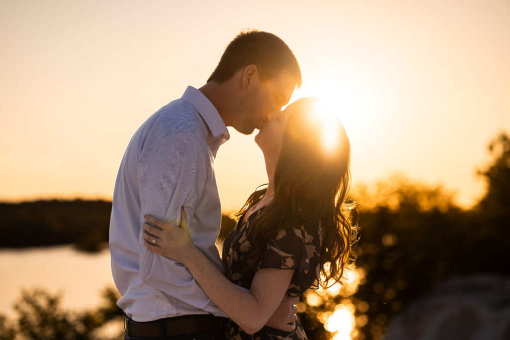 An engagement couple shares a kiss in silhouette during golden hour, standing close together beside a lake with the setting sun glowing directly between their faces. Warm sunlight creates a halo around the woman’s hair while the surrounding trees and water blur into sparkling gold highlights behind them. The scene feels quiet and cinematic, emphasizing their connection against the dramatic sunset backdrop.