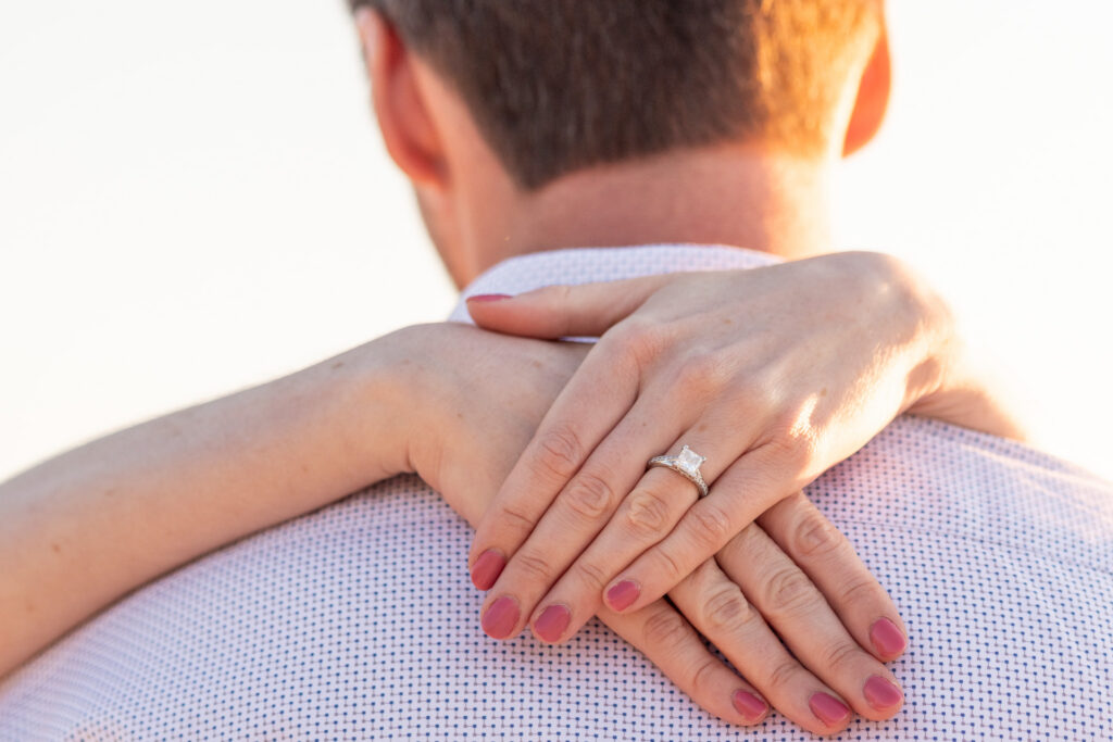 A detailed close-up of an engagement ring resting on a woman’s crossed hands draped over her fiancé’s shoulders. The square-cut diamond ring catches the warm evening light while her soft pink nails and freckles are sharply in focus. The man faces away from the camera in a white patterned dress shirt, and the bright sunset light creates a creamy golden glow around the edges of the image.