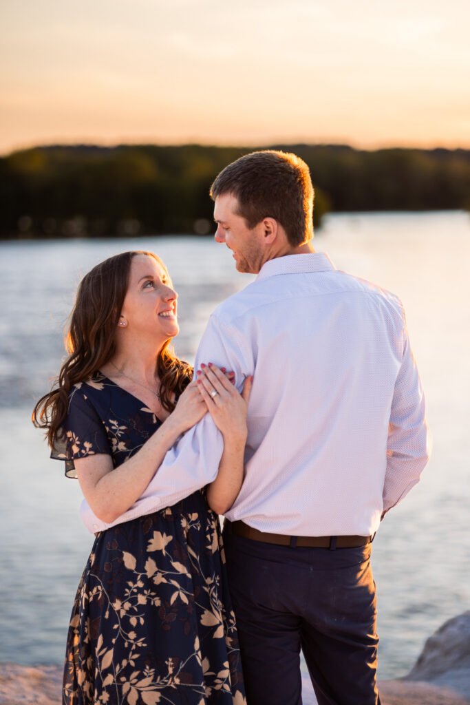 A joyful woman gazes up at her fiancé during a White Cliffs of Conoy engagement session while standing wrapped in his arms beside the water at sunset. Her engagement ring rests visibly against his shoulder as the warm backlight outlines her hair and illuminates the delicate floral pattern of her navy dress. The calm river and distant wooded shoreline blur softly behind them.