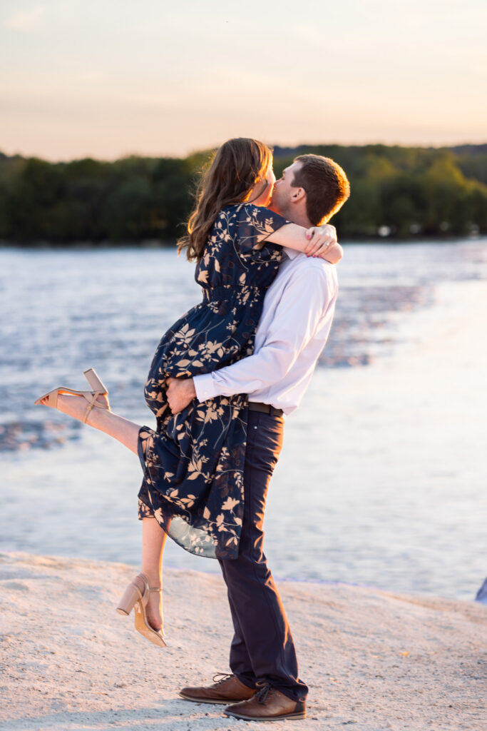 A man lifts his fiancée into the air during a White Cliffs of Conoy engagement session while she wraps both arms around his shoulders and bends one leg playfully behind her. The couple is backlit by warm golden sunset light reflecting off the river beside them, creating a soft glow around their faces and clothing. The pale rocky overlook and calm water create a peaceful, romantic setting.