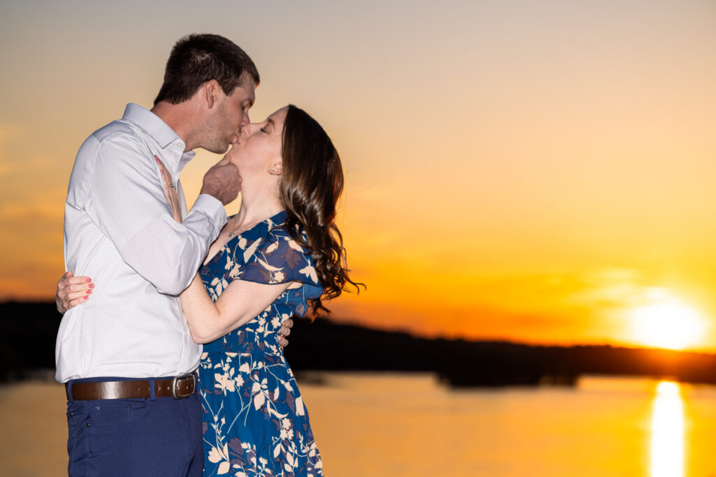 An engaged couple kisses during sunset at a White Cliffs of Conoy engagement session while standing closely together beside the glowing river overlook. The man gently tilts the woman’s chin upward as they embrace, her arm wrapped around his waist and her floral blue dress catching the warm evening light. Behind them, the sky fades from deep orange to soft gold as the sun sinks low over the water, creating a bright reflection across the calm river and silhouetted tree line in the distance.