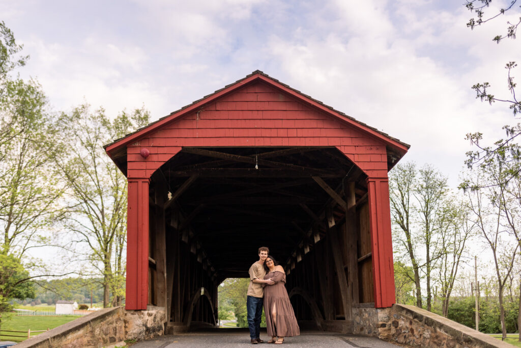 Portrait of Madi and Connor standing together beneath the iconic red covered bridge at Historic Poole Forge in Narvon, Pennsylvania, surrounded by weathered wood, stone walls, and soft evening light.