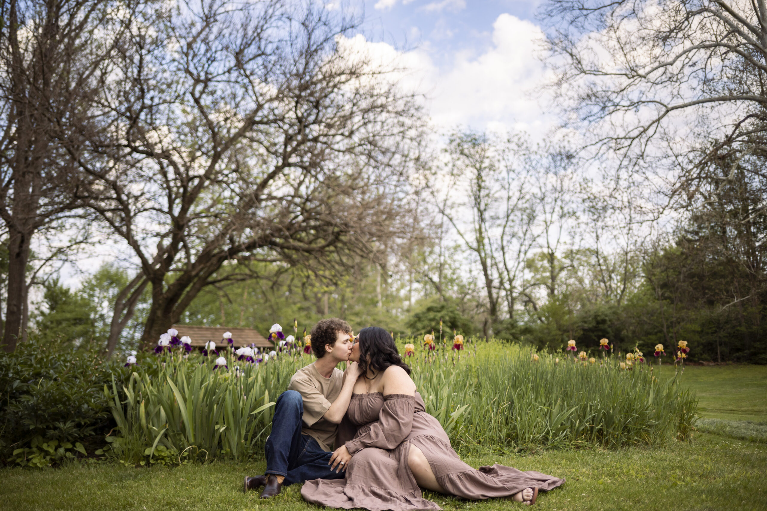 Madi and Connor sitting together in a blooming iris garden during their Historic Poole Forge Engagement Session, sharing a kiss beneath twisting trees and colorful spring flowers near the creekside property.