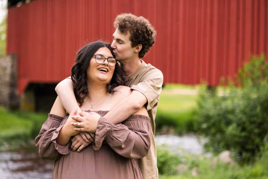Connor wrapping his arms around Madi in front of the red covered bridge during their Historic Poole Forge Engagement Session, while she laughs with her head tilted back in a relaxed and joyful moment.