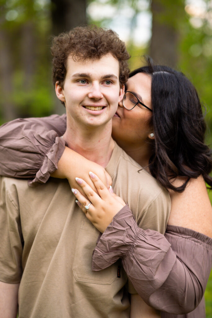 Close-up portrait from their Historic Poole Forge Engagement Session of Madi wrapping her arms around Connor from behind while kissing his cheek, her engagement ring and soft mauve sleeves visible against the blurred green spring background.