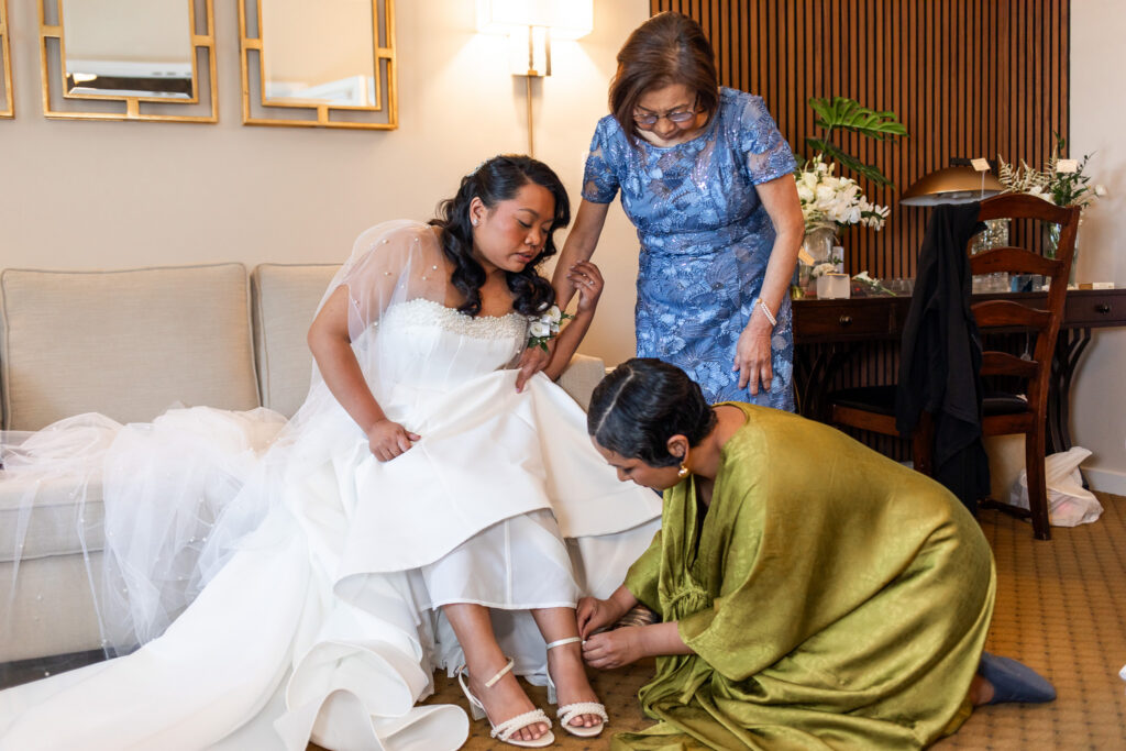 Bride sits gracefully on a cream sofa while family members fasten her pearl-detailed heels, her satin gown spilling dramatically across the floor during getting ready at their Princeton wedding venue.