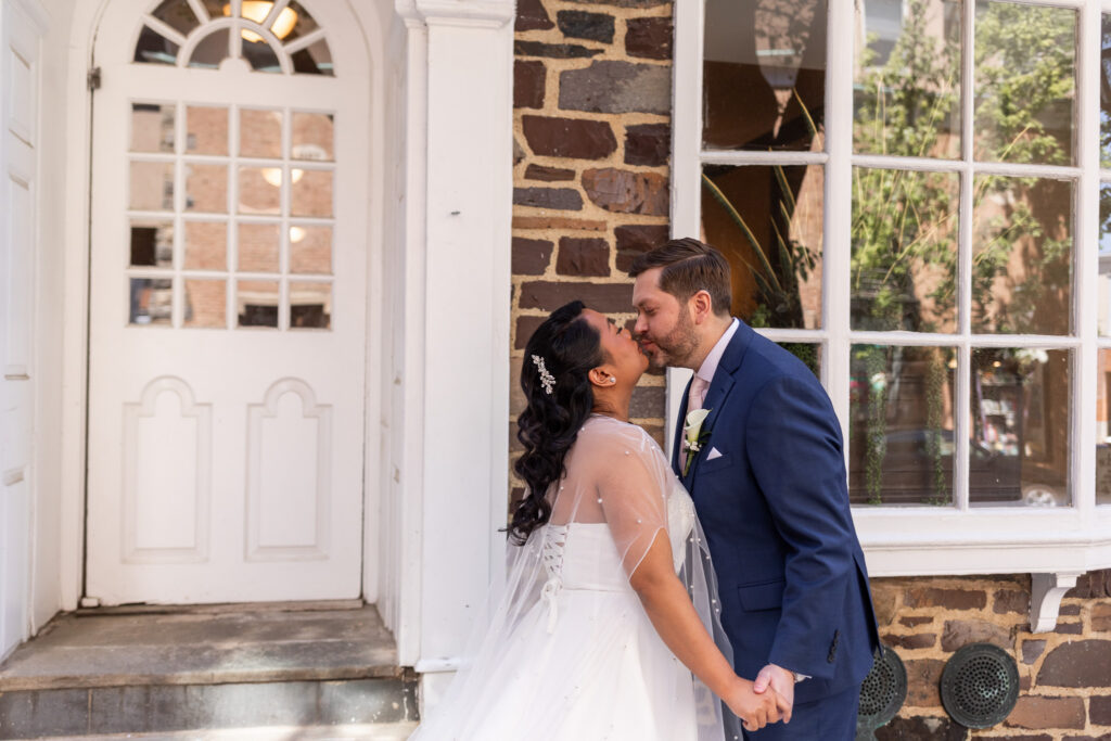 Bride and groom lean in for a kiss beside a white doorway and textured stone wall, sunlight illuminating the bride’s flowing veil during portraits connected to their Princeton wedding venue celebration.
