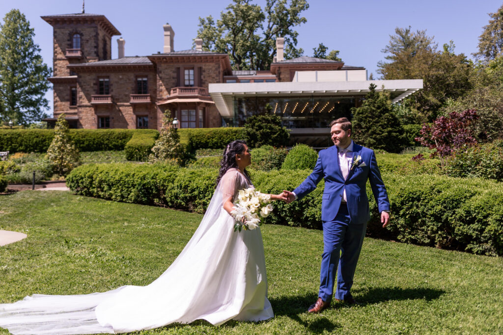 Bride and groom walk hand in hand across the manicured gardens near Prospect House, the bride’s veil flowing behind her during portraits celebrating their Princeton wedding venue wedding day.