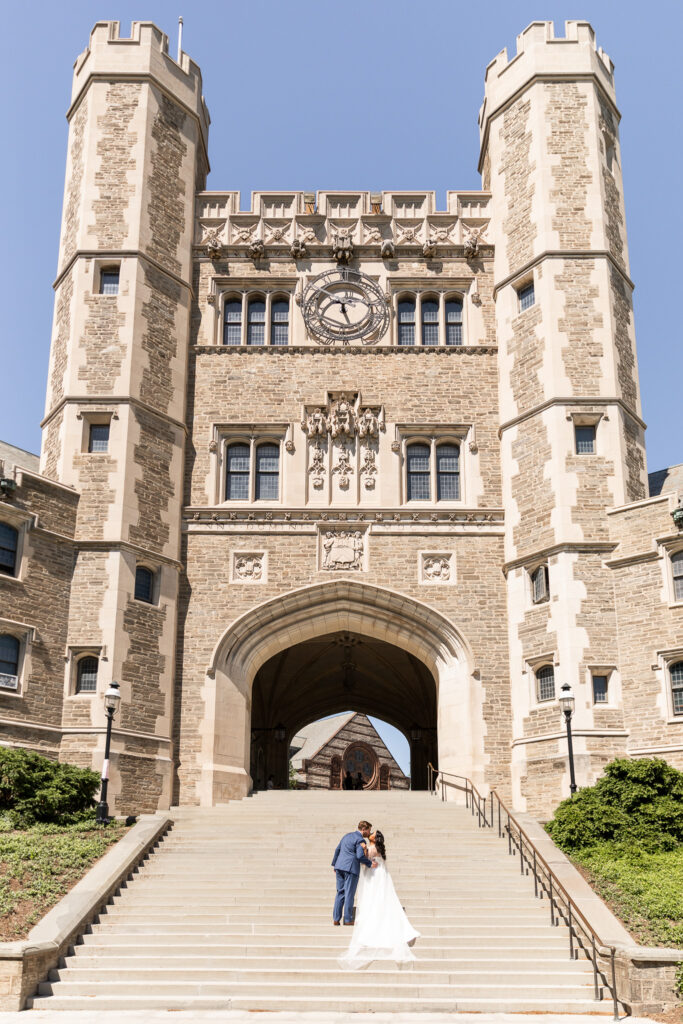 Bride and groom standing together at the bottom of the wide stone staircase beneath Blair Arch, framed by the towering Gothic architecture and large clock above them, with the bride’s veil trailing behind her in the sunlight during portraits at Princeton University.