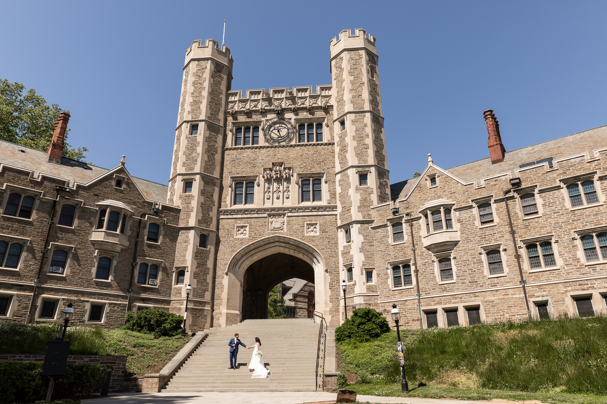 Bride and groom stand hand in hand beneath the towering stone archways of Princeton University, using the historic architecture surrounding their Princeton wedding venue for dramatic wedding portraits beneath a bright blue sky.