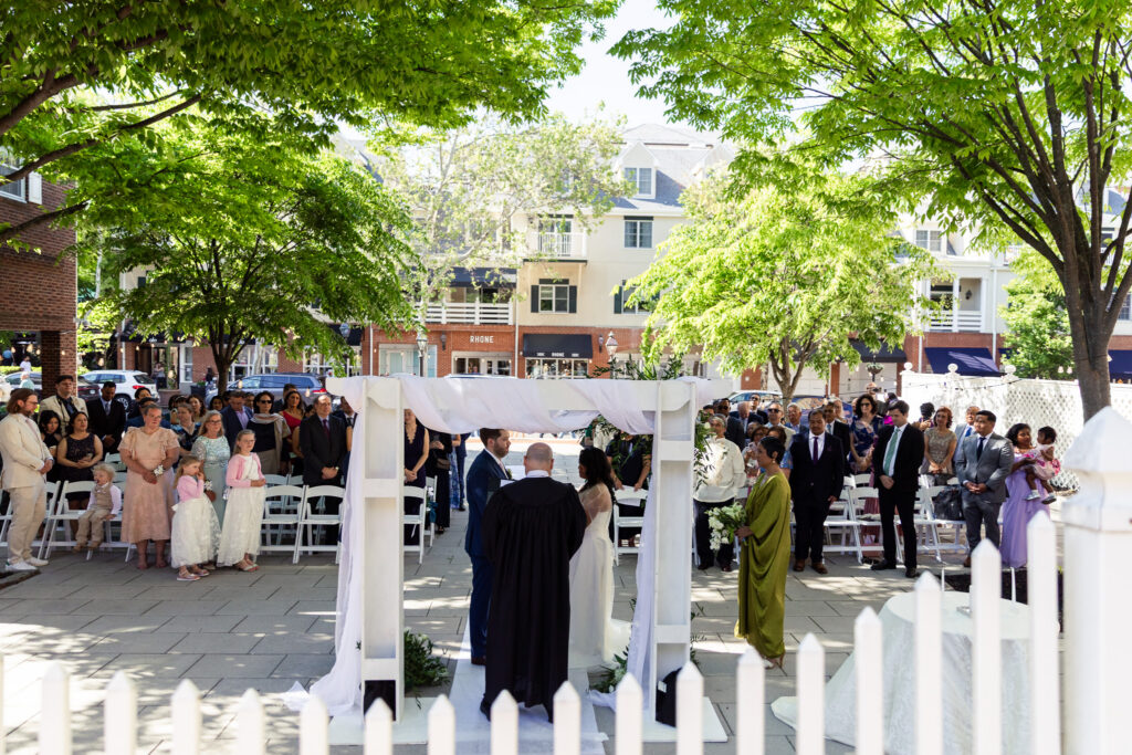Wide ceremony photograph showing guests seated beneath leafy green trees while bride and groom exchange vows under a white draped structure outside their Princeton wedding venue in downtown Princeton.
