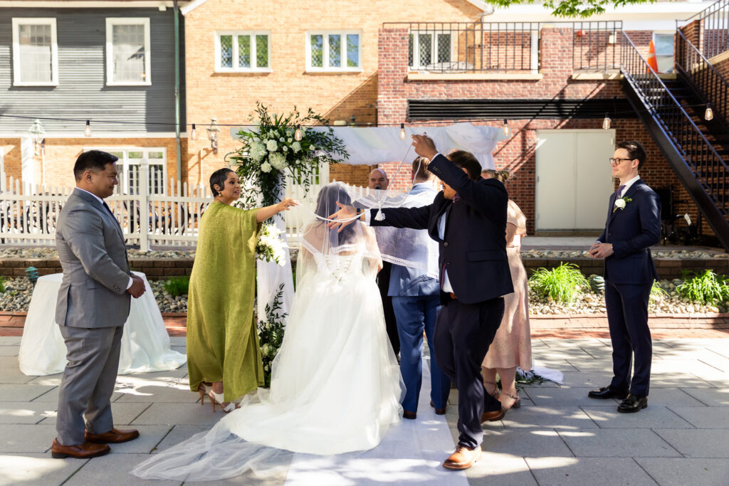 Bride and groom stand beneath a white draped ceremony arbor during an outdoor wedding ceremony as family members carefully lift a traditional veil over the bride’s head. White floral arrangements frame the aisle while guests watch from surrounding chairs in a sunny brick courtyard.