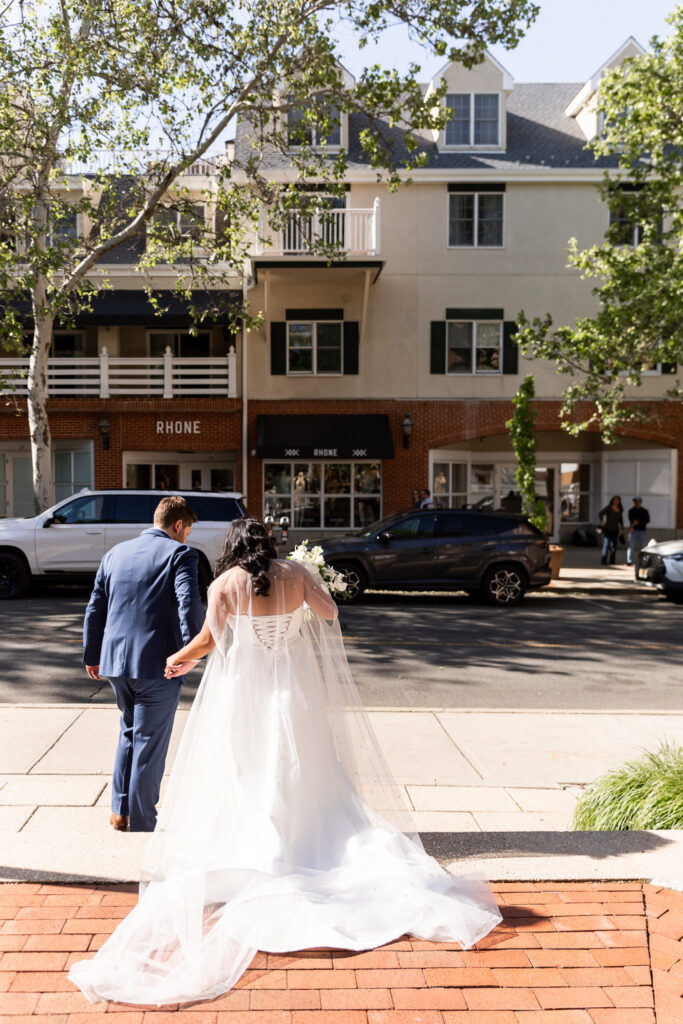 Bride and groom walk hand in hand across a brick sidewalk after their ceremony, the bride’s long veil and train flowing behind her as they cross a quiet street lined with upscale storefronts and tree-covered sidewalks.