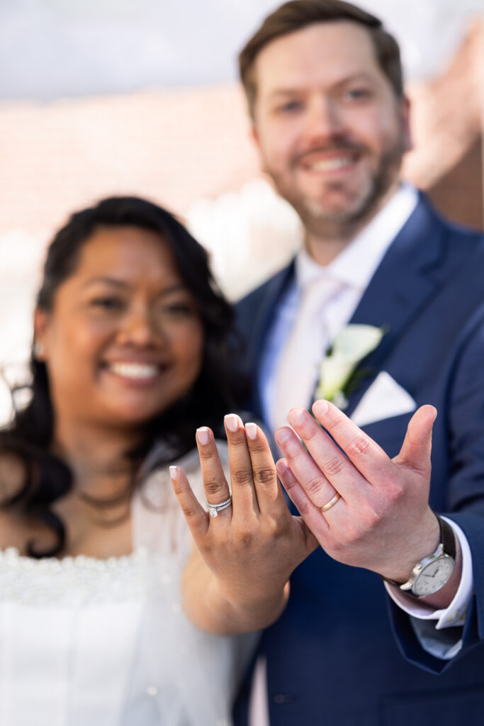 Close up wedding portrait of the bride and groom holding their hands forward to show off their wedding rings, with the bride’s diamond engagement ring and the groom’s gold wedding band in sharp focus while their smiling faces blur softly in the background.