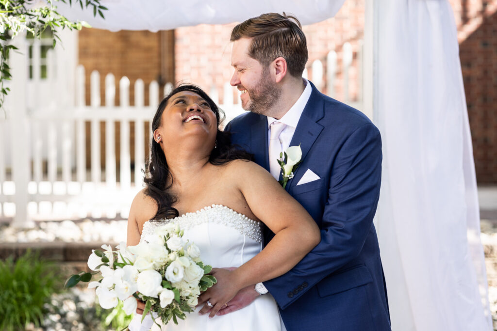 Groom wraps his arms around the bride beneath a white draped ceremony arbor while the bride leans back laughing, holding a bouquet of white flowers during outdoor wedding portraits.