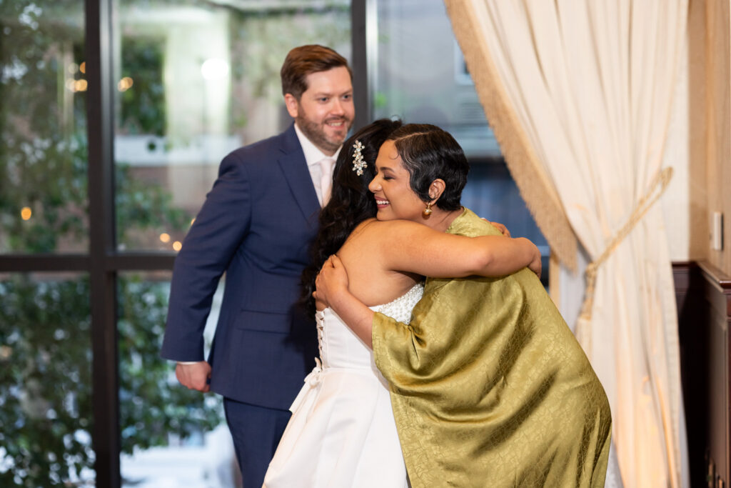 Bride embraces a woman in a shimmering olive green shawl during an emotional reception moment while the groom smiles warmly nearby inside a softly lit ballroom reception space.