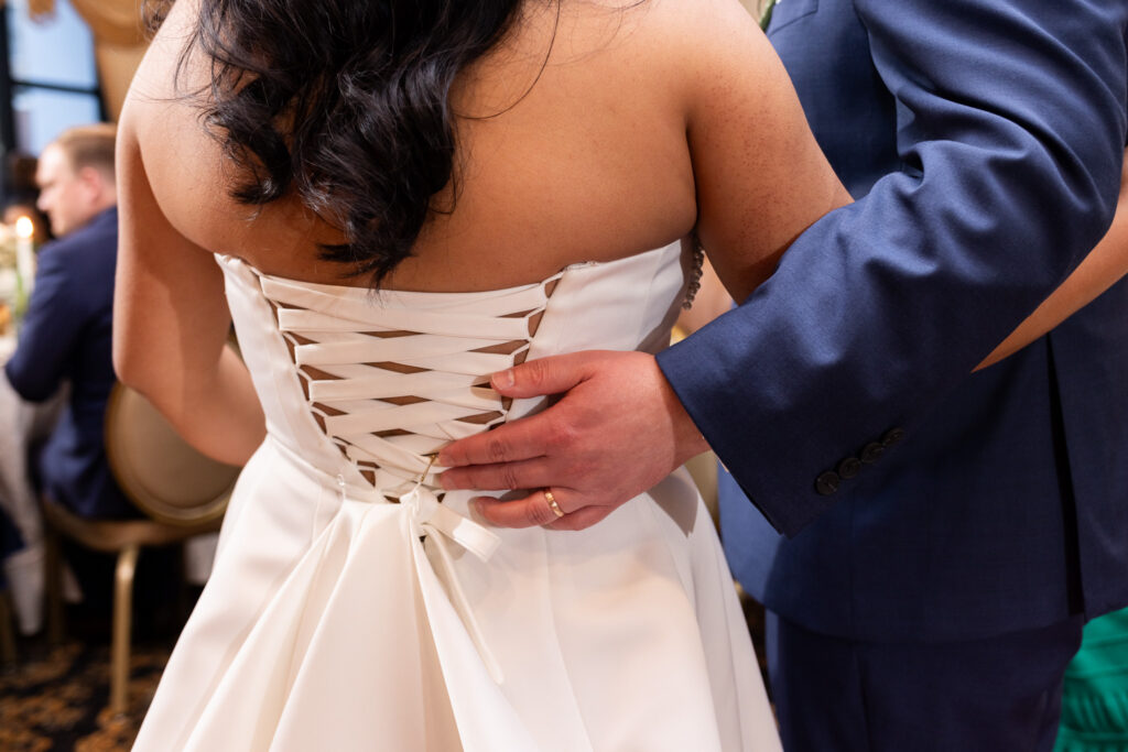 Close up detail of the back of the bride’s wedding dress showing corset-style ribbon lacing as the groom rests his hand gently against her waist during the reception dance floor celebration.