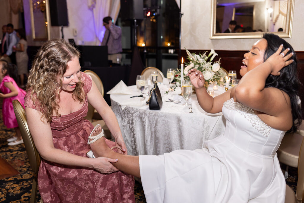 Bride sits laughing during the wedding reception while another guest helps remove her shoes beside a reception table covered in candles and floral arrangements as dancing guests fill the background.