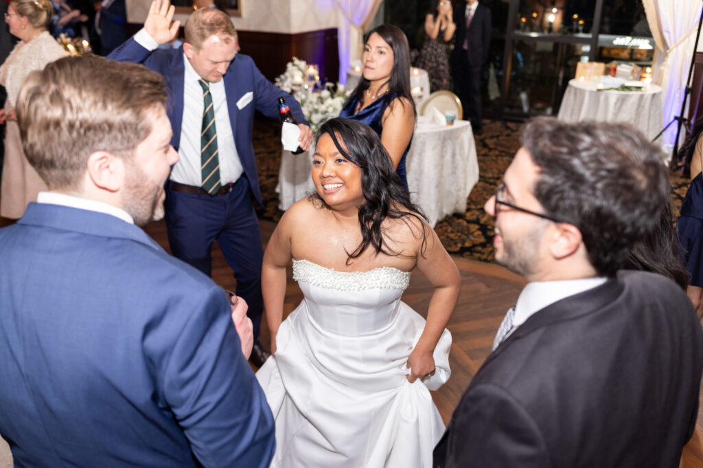Bride smiles brightly while dancing and talking with wedding guests on a crowded reception dance floor filled with movement, formal attire, and colorful evening lighting.