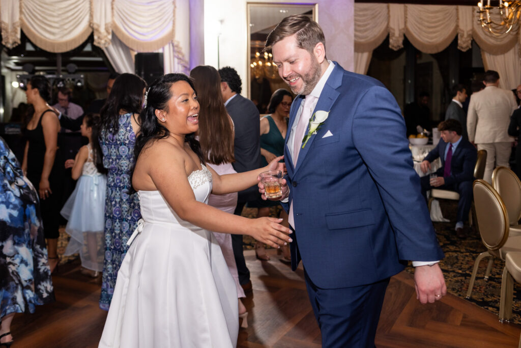Bride and groom laugh together while dancing during the wedding reception, surrounded by guests on a lively ballroom dance floor with elegant drapery and candlelit tables in the background.