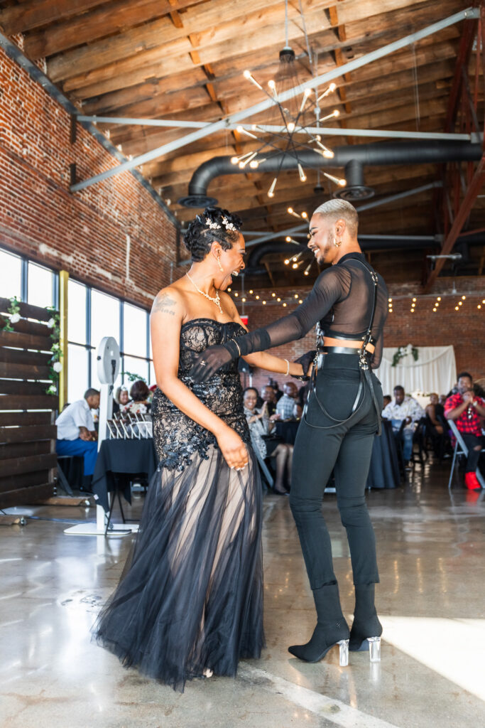 A non-traditional groom shares a dance with his mother during an LGBTQ wedding reception in an industrial-style reception space with exposed brick walls and modern chandeliers. Mom wears a dramatic black lace gown while the groom wears a fashion-forward black outfit with sheer sleeves and high-heeled boots. Moments like these reflect how LGBTQ-friendly wedding vendors help create celebrations where self-expression and individuality are fully embraced.