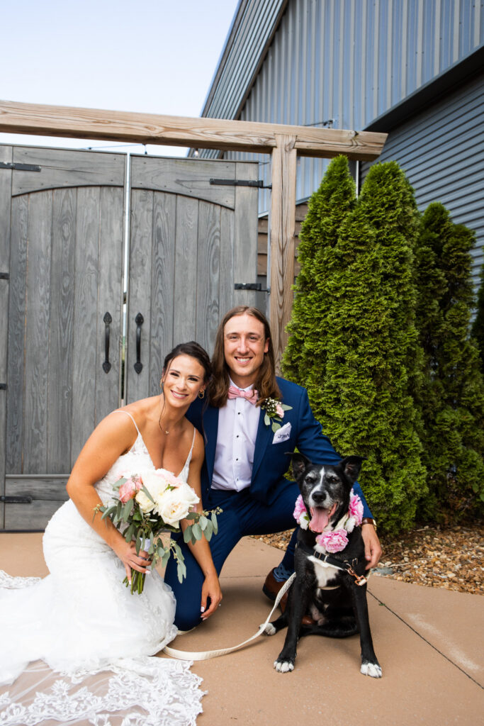 A bride and groom kneel beside their black-and-white dog outside a rustic wedding venue. The dog  ring bearer wears a pink floral collar that coordinates with the wedding flowers while the couple smiles toward the camera during their wedding portraits.