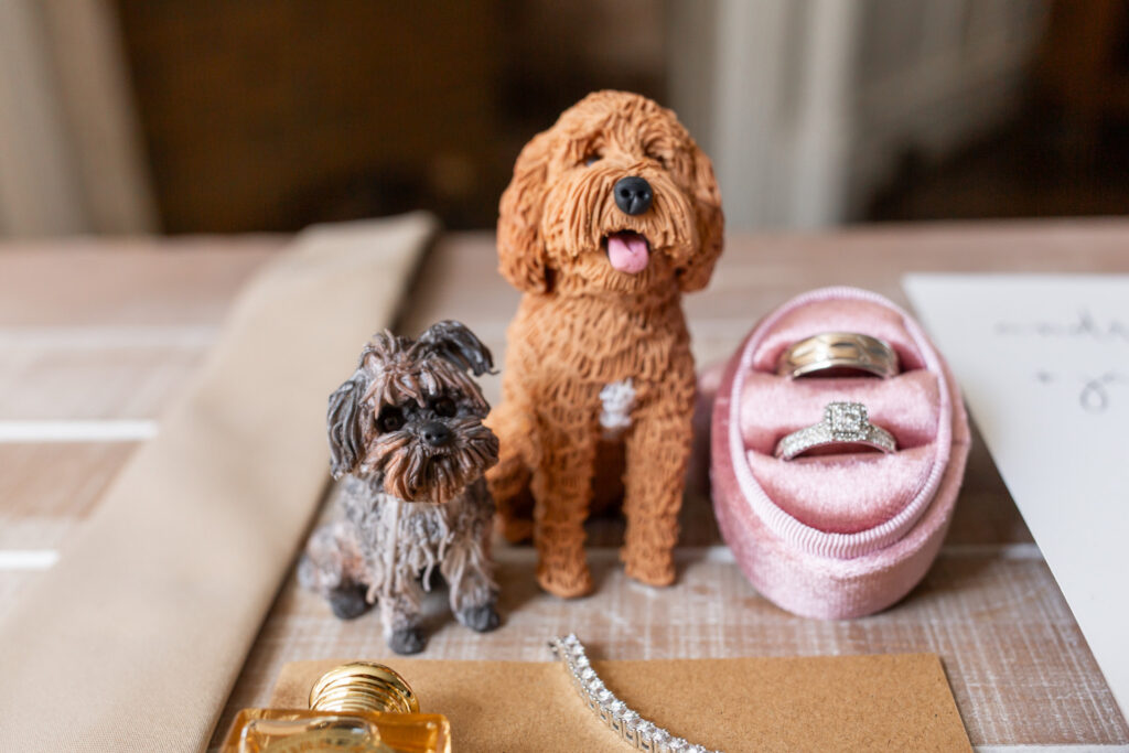 Wedding detail photograph featuring two custom dog figurines positioned beside the couple's wedding rings and jewelry. The figurines resemble the couple's pets and sit among carefully styled wedding details, offering a creative way to include dogs when they cannot attend the celebration.