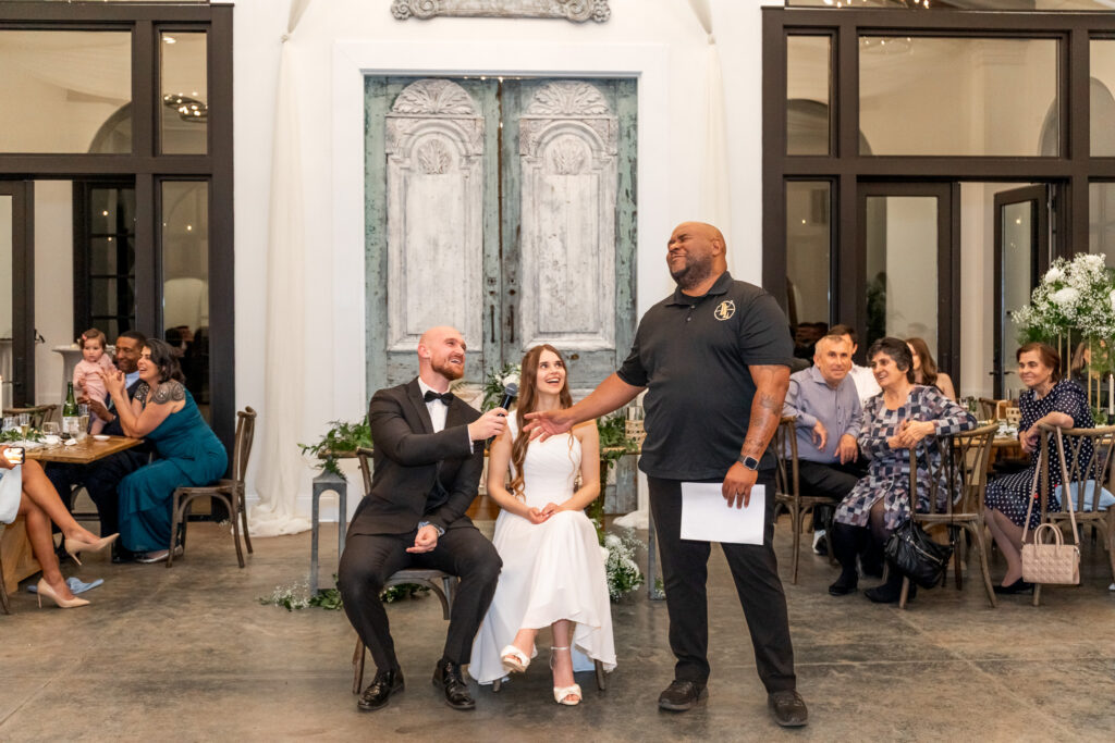 A DJ stands with a microphone during a reception game while the couple laughs from their seats and guests watch from surrounding tables.