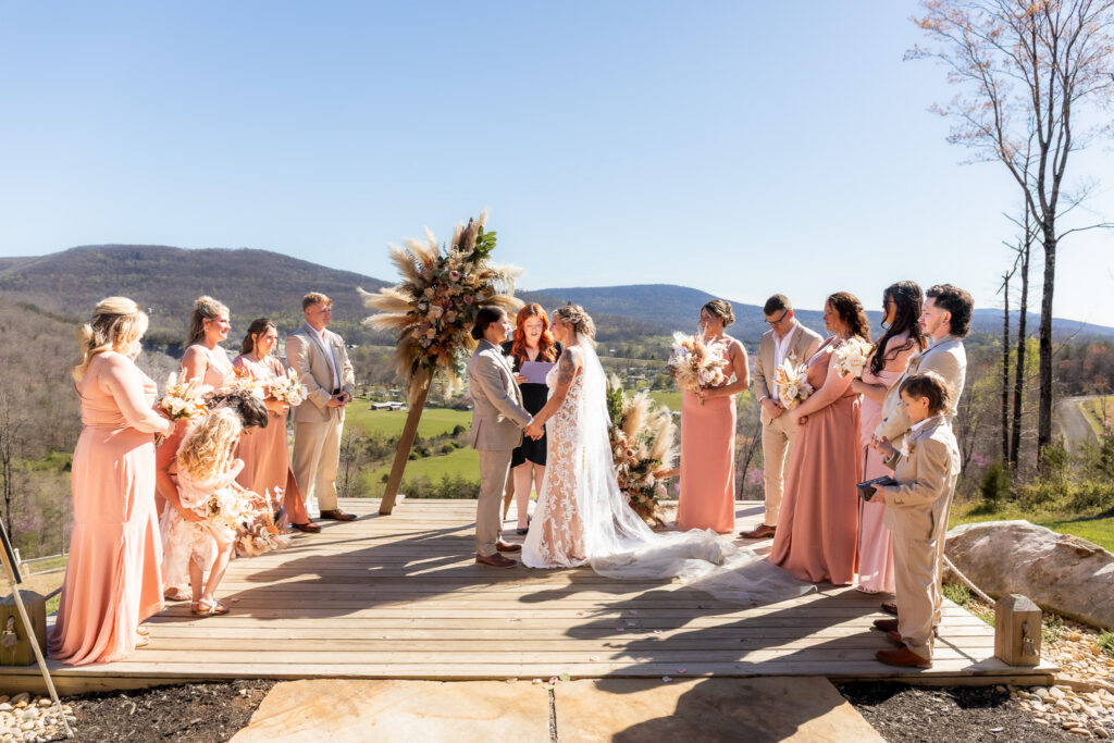 A couple stands at the center of an outdoor ceremony overlooking mountain views, surrounded by their wedding party in coordinating blush and tan attire beneath a floral arbor.