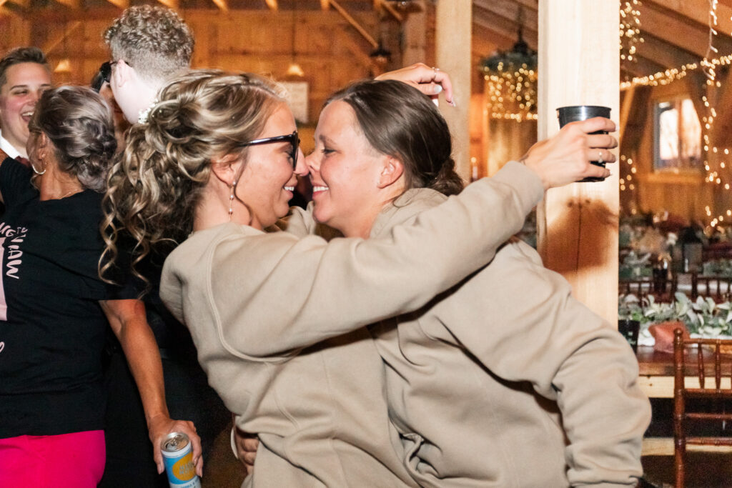 Two brides in matching tan hoodies share a playful embrace on the dance floor, smiling nose-to-nose beneath warm string lights during their LGBTQ wedding reception.