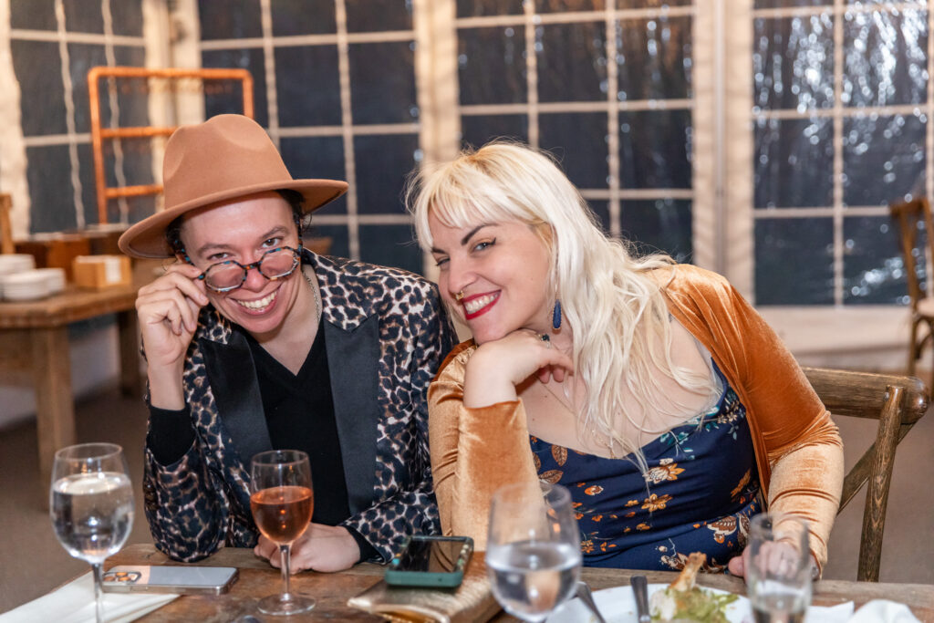 Two wedding guests sit together at a reception table, smiling directly at the camera. One wears a leopard-print jacket and wide-brim hat while the other wears a floral dress and gold velvet cardigan, showcasing the individuality often celebrated by inclusive wedding vendors.