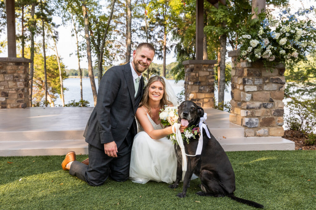 A newly married couple kneels beside their black Labrador dog ring bearer outside a lakeside ceremony pavilion. The dog wears floral accents and ribbon adornments while the couple smiles toward the camera with the water and stone structure visible behind them.