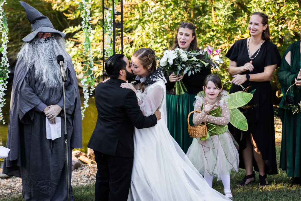 A couple shares their first kiss after being pronounced married during an outdoor wedding ceremony officiated by a loved one dressed as Gandalf the grey, while wedding party members and a flower fairy celebrate nearby.