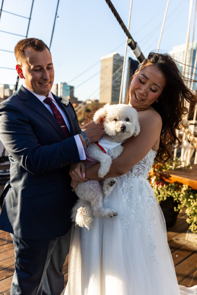 A newlywed couple stands together on a historic ship deck while holding their small white dog. Warm evening sunlight illuminates the scene as the dog looks directly toward the camera, becoming part of the couple's wedding portraits.