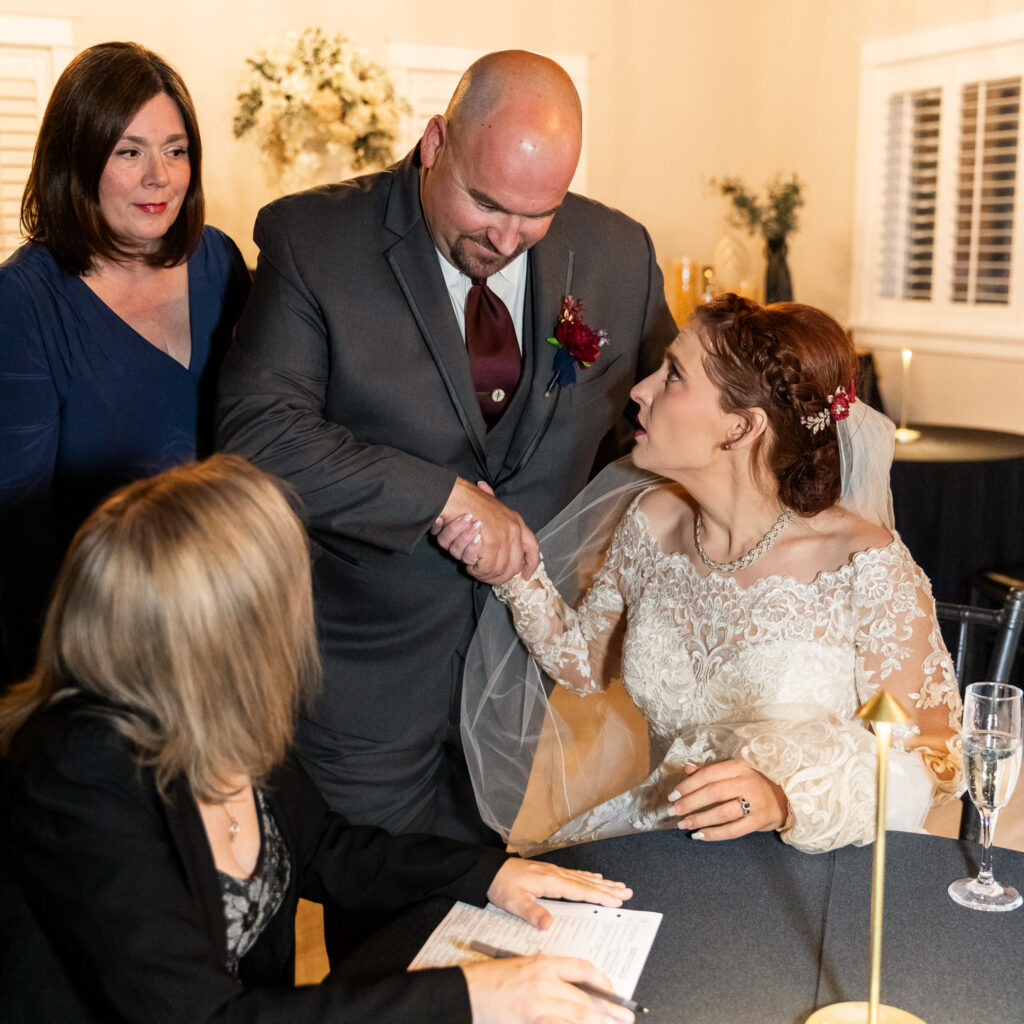 Newlywed couple share a jokingly serious handshake over their license paperwork during a wedding reception while witnesses and their officiant gather around the table, documenting the legal moment that officially completes the marriage.