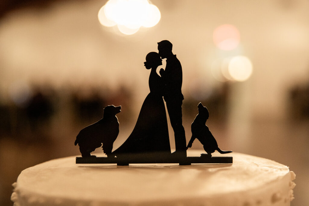 Close-up photograph of a wedding cake topper silhouetted against soft reception lights. The design features a bride and groom standing together with two dogs, creating a meaningful tribute to beloved pets who are part of the couple's story.