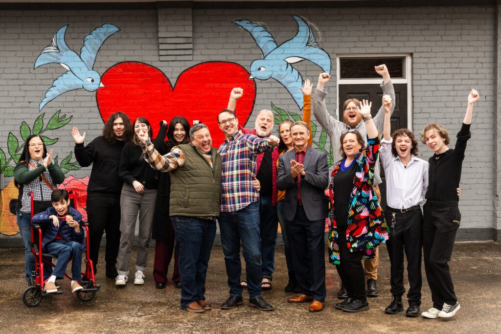 A large chosen family group gathers in front of a colorful mural featuring a red heart and blue birds, cheering with raised hands and joyful expressions after an LGBTQ wedding celebration. Many LGBT friendly wedding vendors recognize that family can be defined by love and support rather than tradition alone