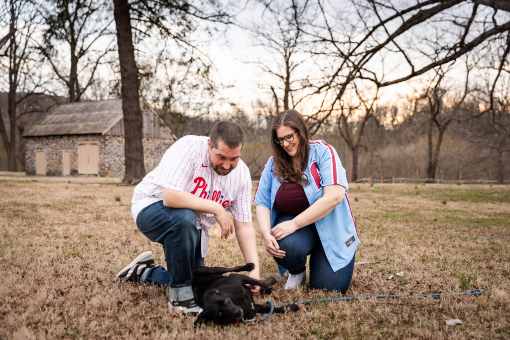 A couple wearing Philadelphia Phillies jerseys kneels in a grassy field during their engagement session while their black dog rolls onto its back for belly rubs. The playful moment captures the dog's personality as the couple laughs and interacts with their beloved pet in front of a historic stone building.